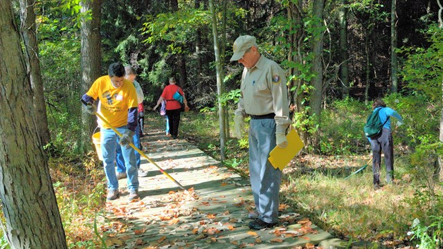 Man in khaki volunteer uniform stands on a bridge in a forest near other people holding rakes.