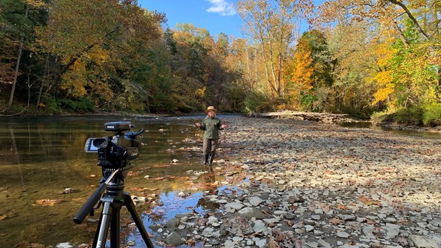 Uniformed ranger stands in front of a video camera in water near the edge of a river.