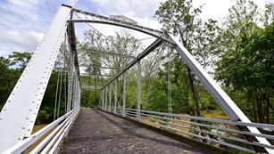 View down a rusting white metal truss bridge with a wooden floor spanning a muddy river.