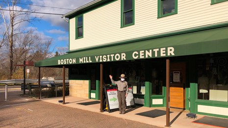 A masked, female ranger waves hello in front of a white building with a green awning.