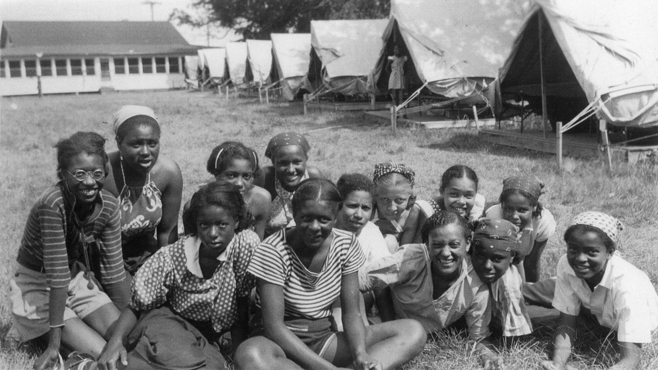13 African American girls sit on a lawn in front of a row of tents.