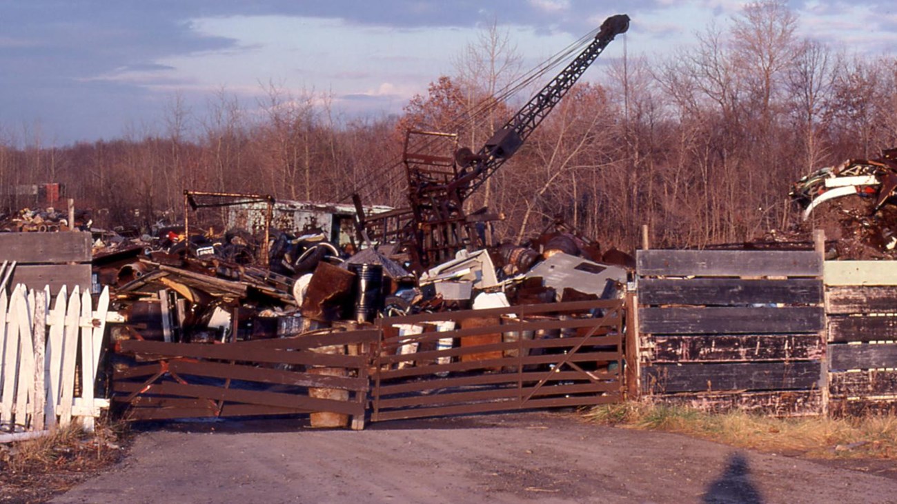Behind a fence of wood and metal is a large industrial waste dump. A crane and pulley reach the sky.