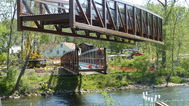 Section of brown, metal bridge suspended over a river as workers wait on another section nearby.