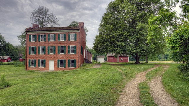 A 3-story brick building with many windows and green shutters with red outbuildings behind it.