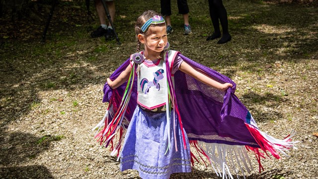 Young girl in a colorful dress stands smiling, her outstretched arms holding her fringed cape open.
