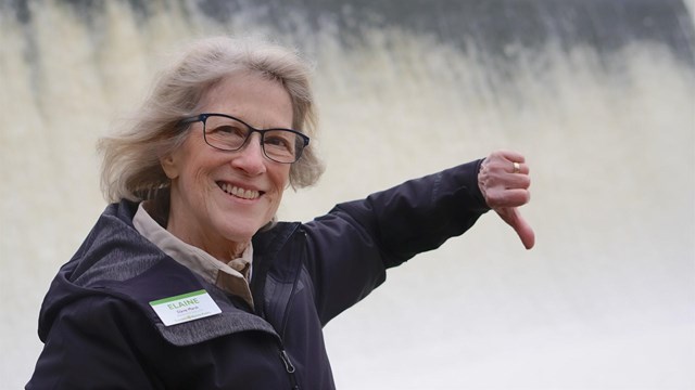Woman with a Summit Metro Parks nametag gives a thumbs down to the wall of rushing water behind her.