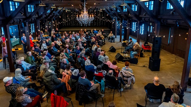 About a hundred people sit in a semi-circle in a large room while two musicians play.