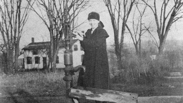 A black-and-white photo of a woman in a long, black dress standing at a water pump.