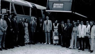A black-and-white photo of twenty people, mostly men dressed in suits; behind them, two tour buses.