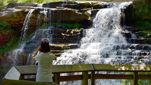 A photographer stands on the lower viewing platform as they observe the roaring waterfall.