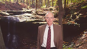 Man with white hair in a tan suit stands in front of a waterfall surrounded by trees.