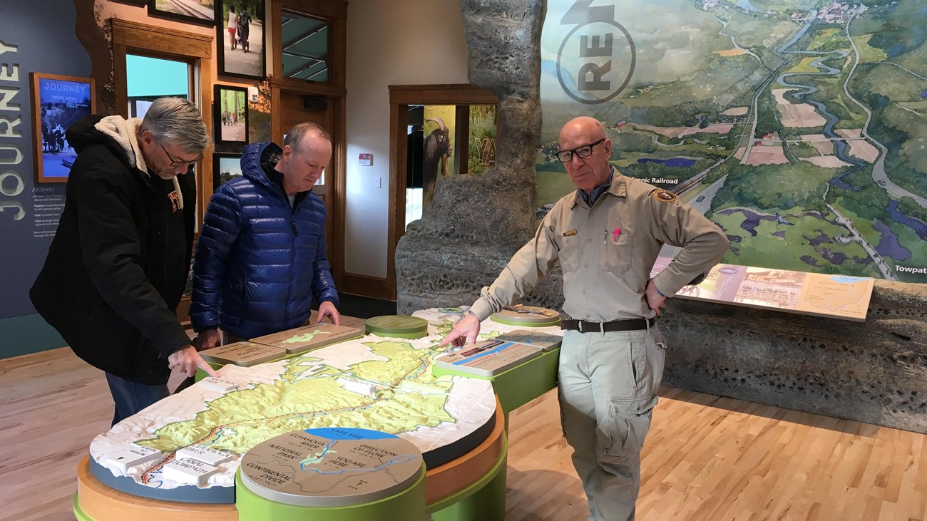 Man in khaki volunteer uniform stands with two others, pointing to a map table; a mural behind them.
