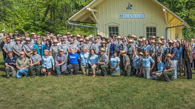 Over 100 mostly uniformed people stand or kneel in rows in front of a small, yellow building.