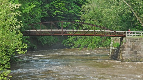 An iron, parabolic pedestrian bridge extends across a rushing river surrounded by trees.
