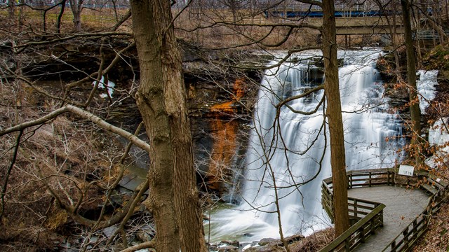 View down through bare trees at a deck overlooking a waterfall cascading into a gorge.