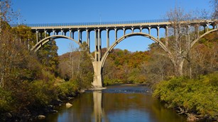 An arched concrete bridge high over a river with orange leaves and a blue sky behind.