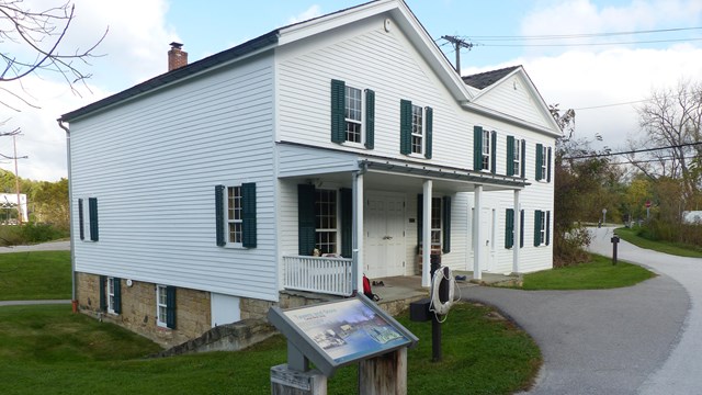 A graphic panel and life ring stand outside a white, two-story historic building with a porch.