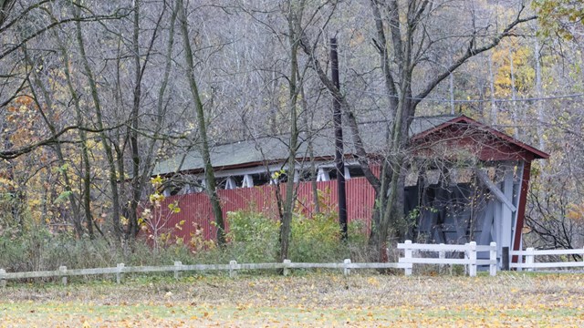 Among the trees, behind a low white rail fence, is a red covered bridge with a white interior.