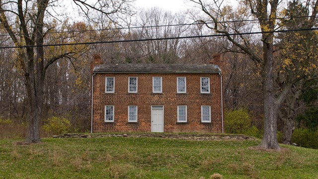 A two-story brick building with rows of white-paned windows, on a hill surrounded by trees.