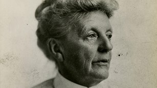 Black-and-white photo: older woman with pale hair in a bun, pleated white shirt pinned at collar.