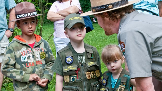A uniformed ranger speaks to three children in green vests covered in Junior Ranger badges