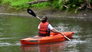 Person wearing an orange life vest sits in an orange kayak on the river, holding a paddle.