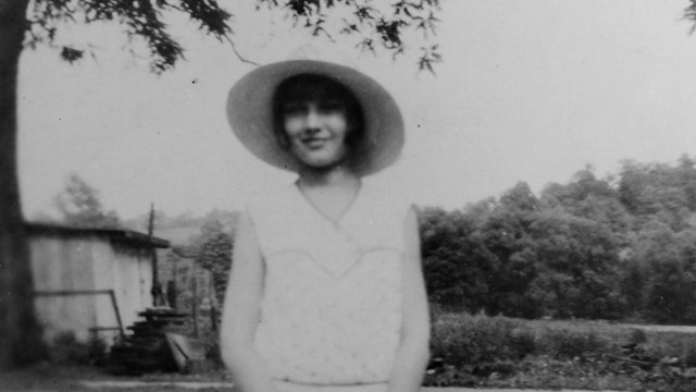 Black-and-white photo of a girl wearing a broad-brimmed hat, a polka-dot dress, and white tights.