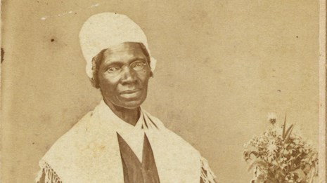 An African American woman sits in long dark dress and white shawl and cap, knitting.