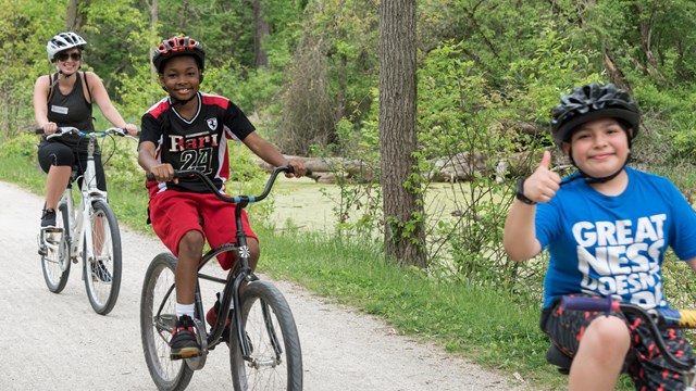 Three children smile and ride bikes down an unpaved path, green trees behind them.