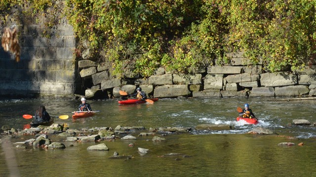 Four people in orange kayaks paddled in a shallow river in front of a stone embankment.