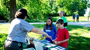 A ranger talks to children and parent at a table outside in front of green trees.