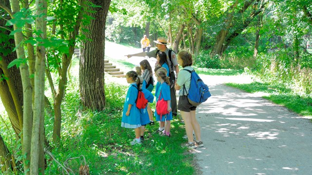 A group of children wearing blue shirts stand with a uniformed ranger who points off to the left.