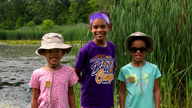 Three young girls with Junior Ranger badges stand in front of a pond.