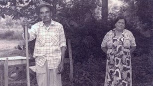 Black and white photo of a couple standing in a yard next to a chicken pen and trees.