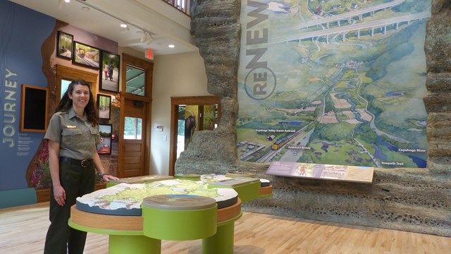 A female uniformed ranger stands indoors next to a map table with a large mural in the background.