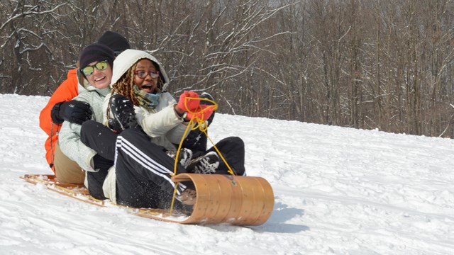 Three people in winter coats and gloves smile as they ride down a snowy hill on a wooden toboggan.