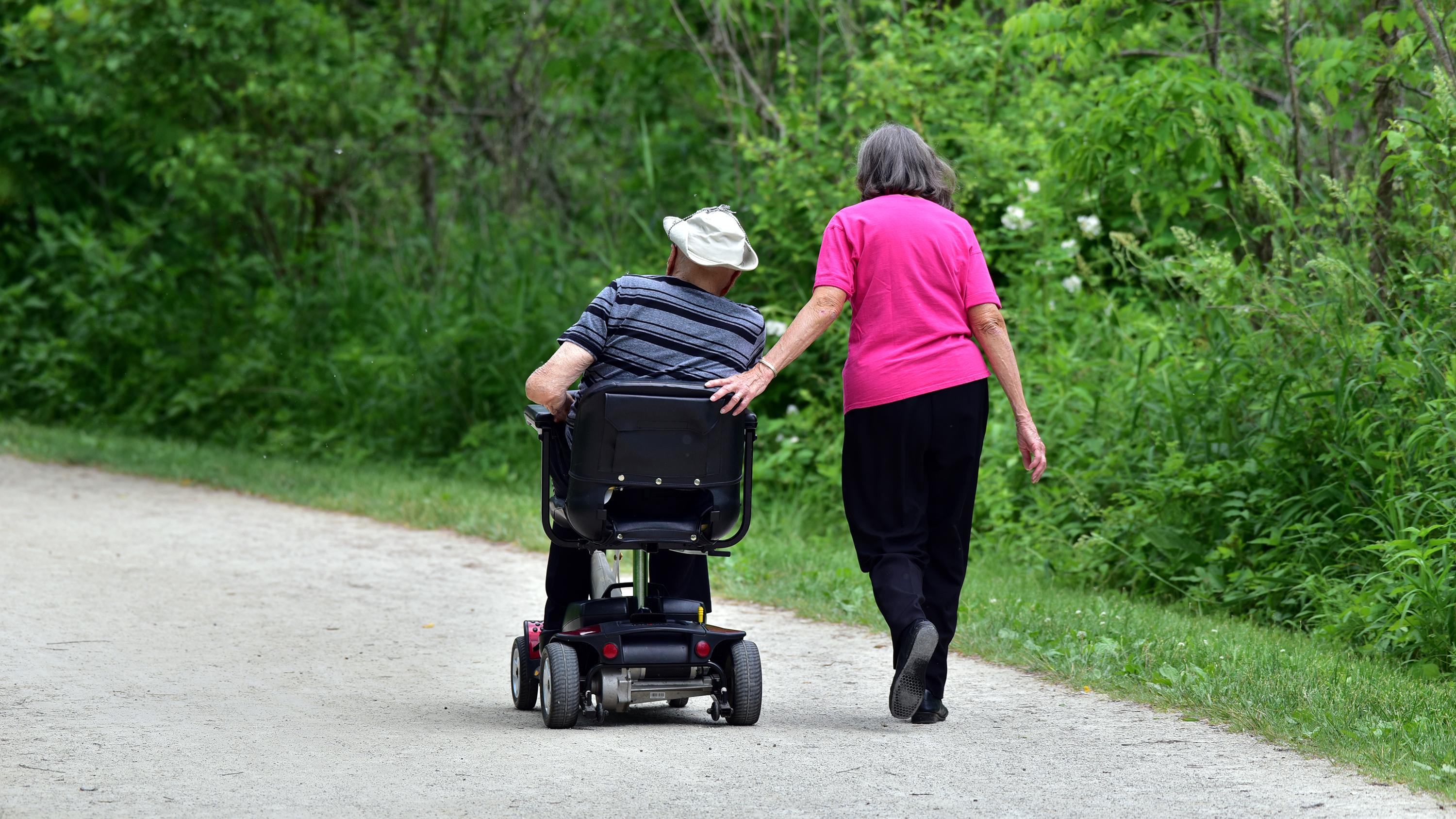 Two people, one walking one in a motorized scooter, on a trail outdoors.