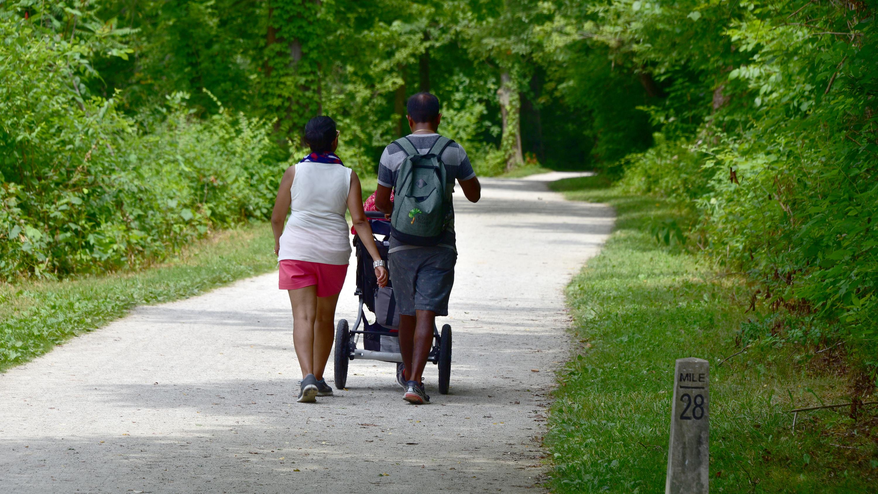 A couple walk with a stroller down a wide, gray path through a green forest.
