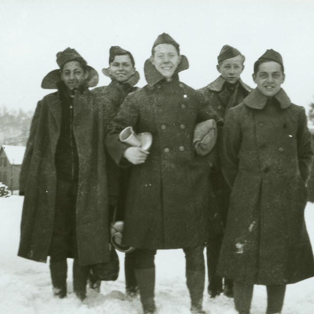 Black and white photo of six smiling young men standing out in the snow, wearing long wool coats.