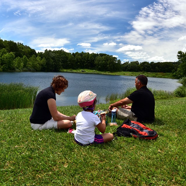 Two adults and a child sit in mowed grass and eat; the child wears a bike helmet.