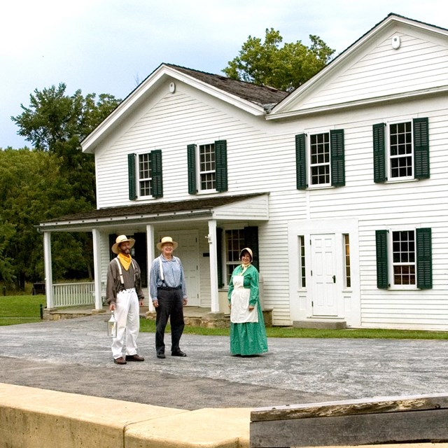 Three people in canal-era dress stand in front of a white building with green shutters.