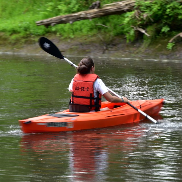 Person wearing an orange life vest sits in an orange kayak on the river, holding a paddle.