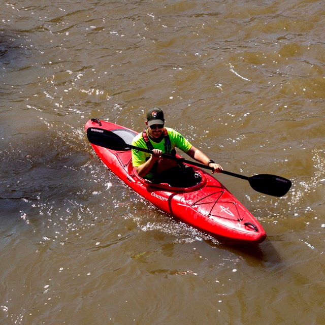 Ranger in bright yellow shirt and red life vest paddles a red boat on brown water.