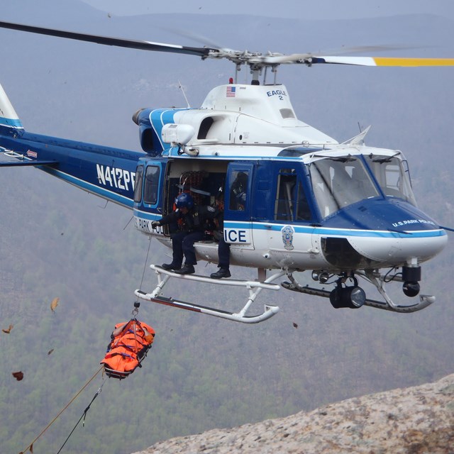 A helicopter kicks up leaves and debris as it hoists a stretcher above a forest