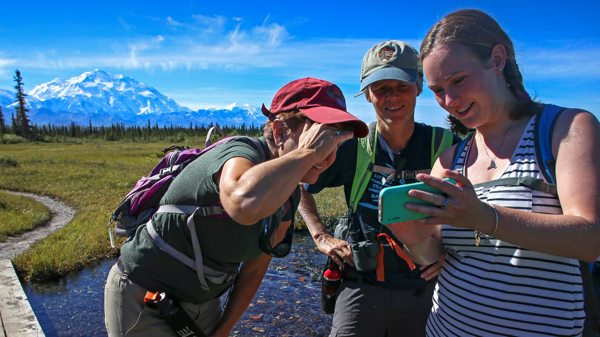 Two hikers and one guide near a creek with a mountain in the background, stopped to look at a pictur