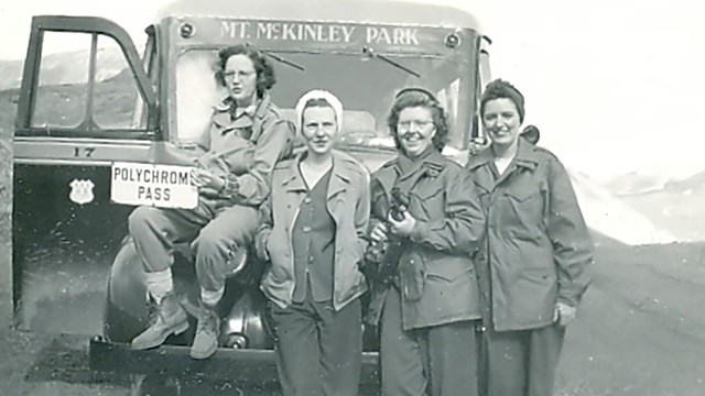 four women standing in front of a bus on a dirt road