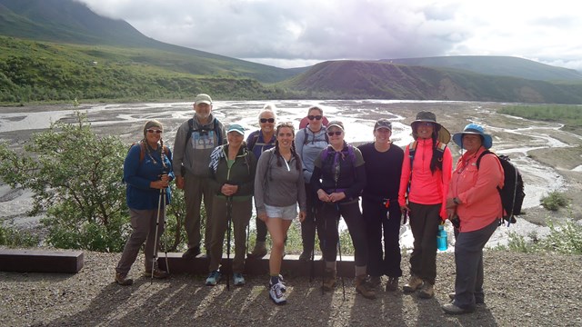 A group of 10 people stand on the edge of a gravel road in front of a river and mountains.