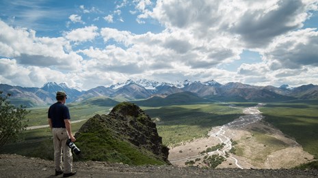 a man standing at a scenic overlook above a wide green plain leading up to distant mountains