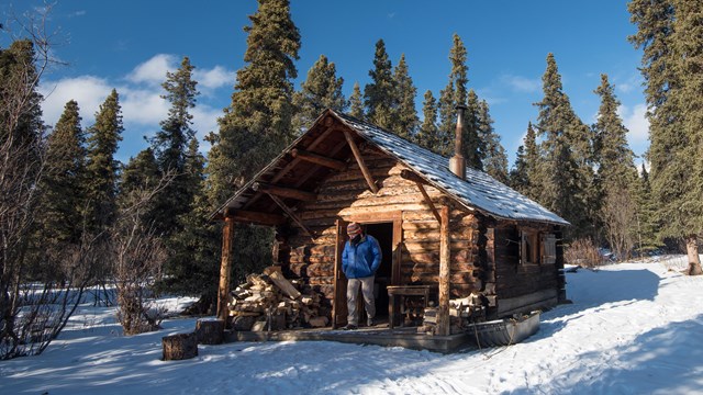 a man stands in the doorway of a small cabin surrounded by snow and spruce trees