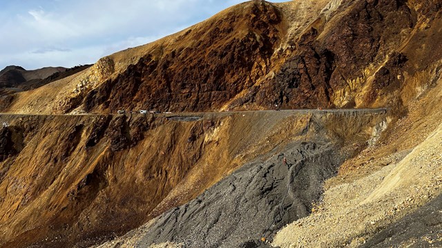 A mountainside of orange and gray rock slumping downhill away from the flat road bed.